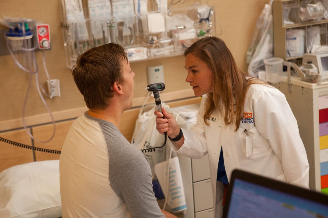 A healthcare professional examines a patient's throat with an otoscope during an urgent care virtual visit in a medical examination room.