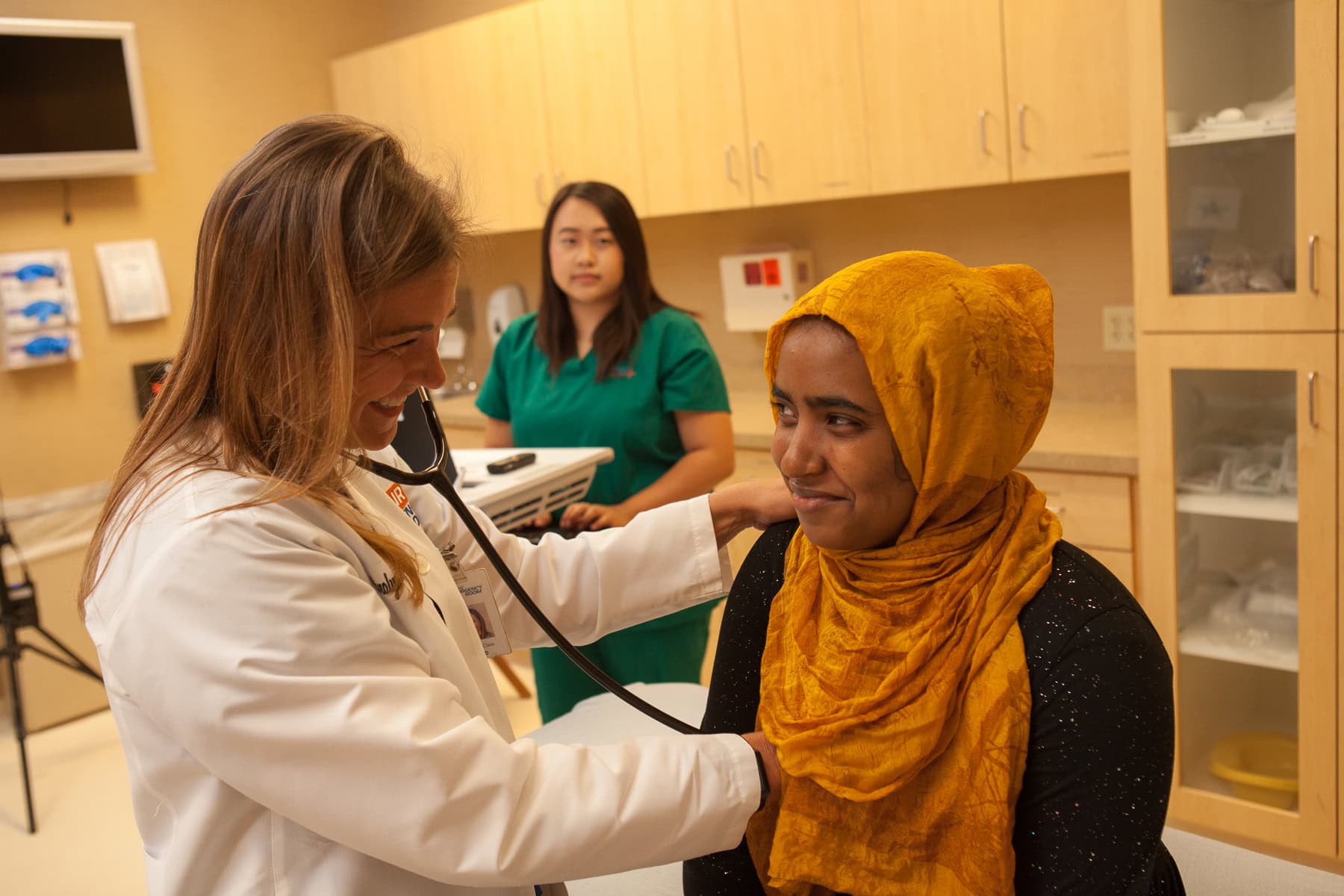 A doctor uses a stethoscope on a woman in a yellow headscarf as a nurse looks on, highlighting the ease of online urgent care visits.