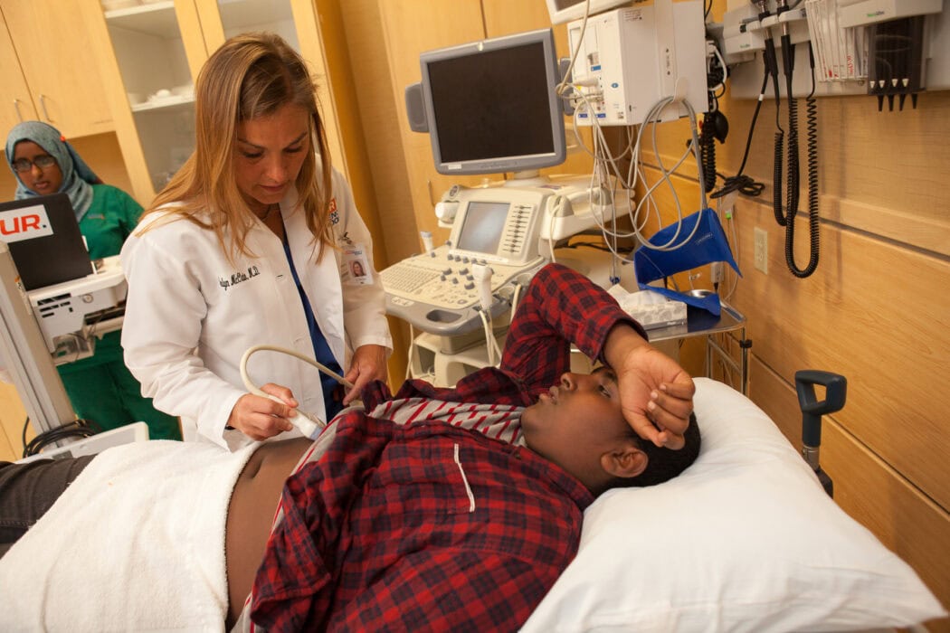A doctor performs an abdominal ultrasound during an urgent care virtual visit in a hospital examination room.