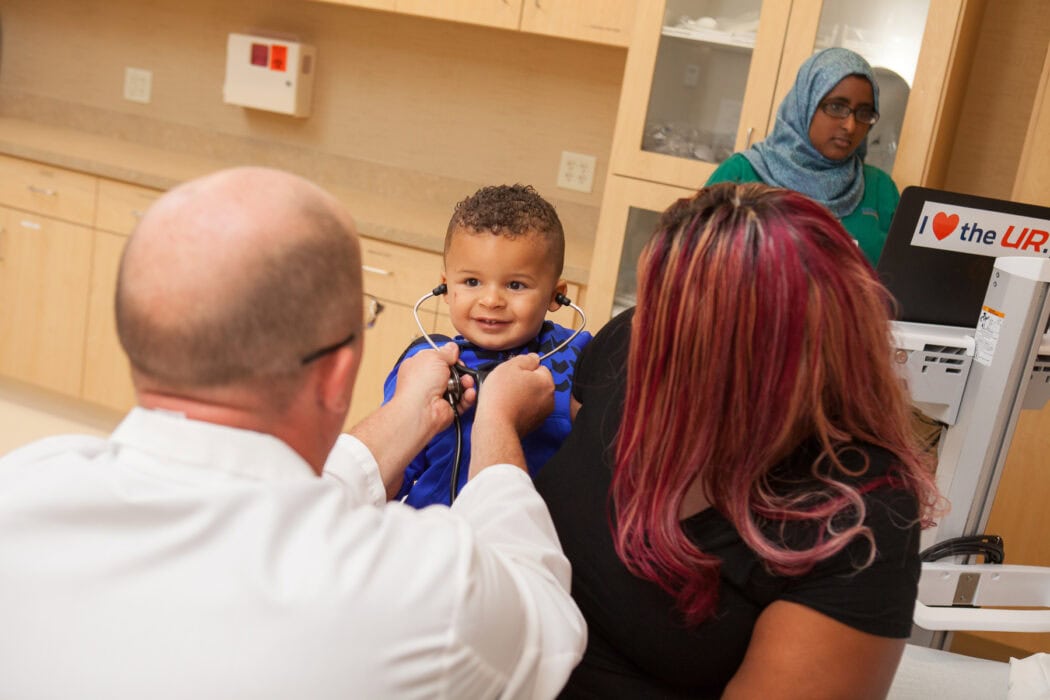 A doctor uses a stethoscope on a smiling toddler in an exam room, highlighting the comfort of an urgent care virtual visit experience.