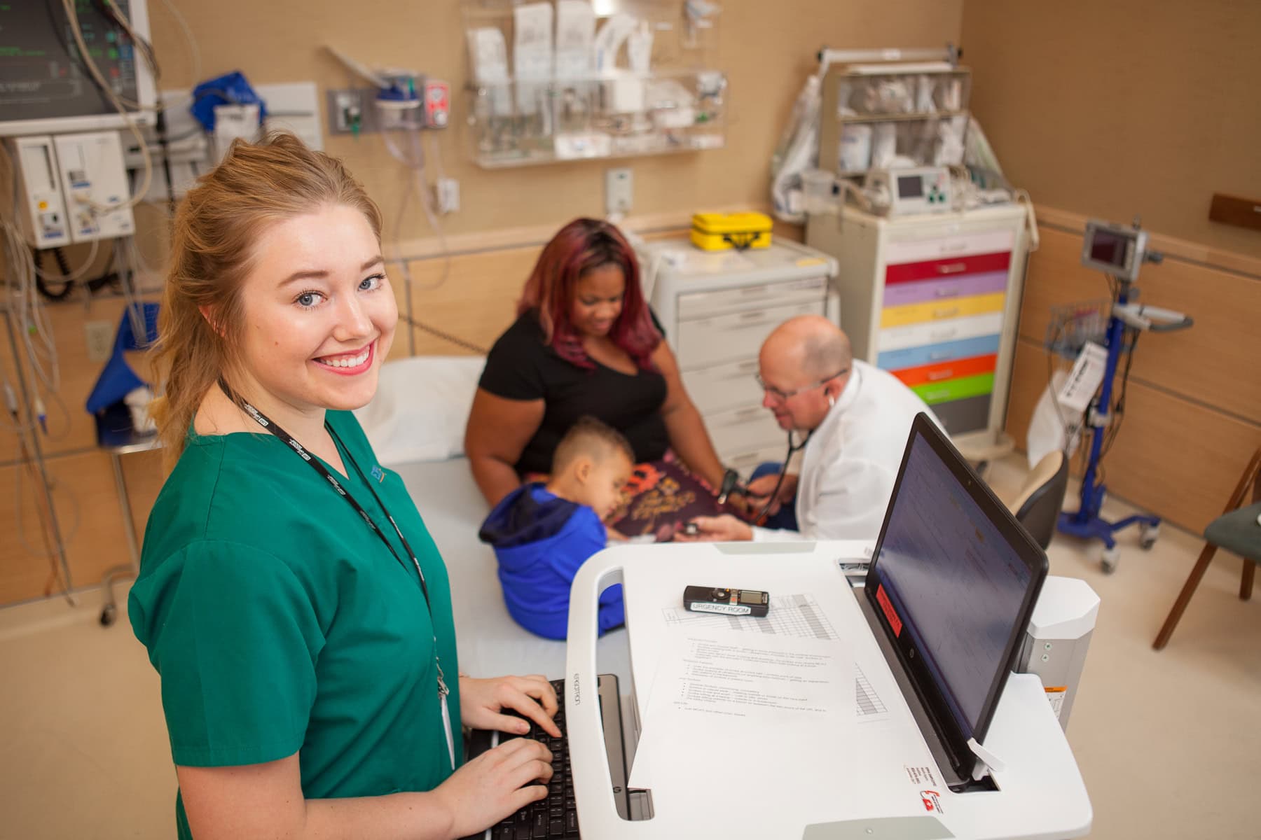 A nurse types at a computer while a doctor examines a child in the urgency room with his mother on a hospital bed.