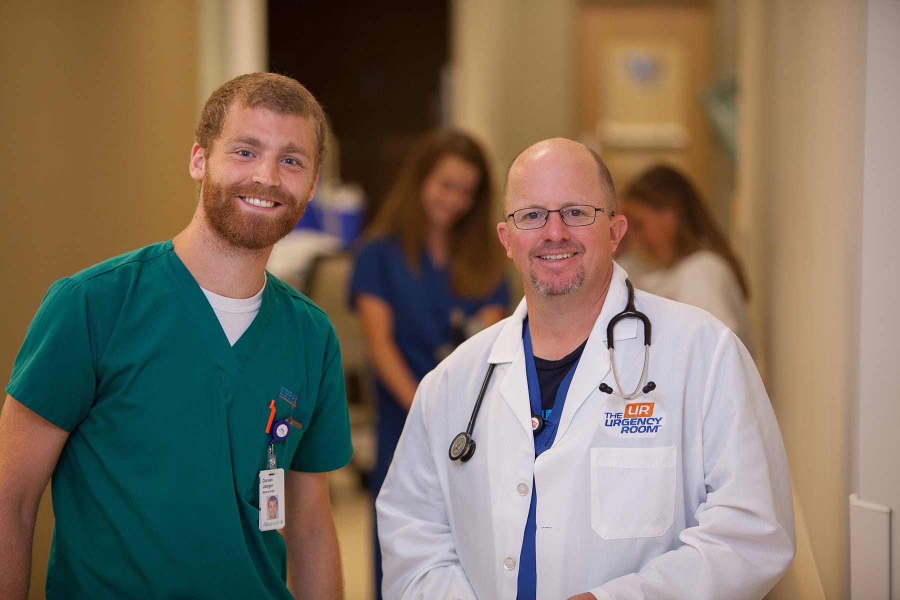 Two healthcare professionals, in green scrubs and a white coat, smile in the urgency room hallway—ready for your urgent care online needs.