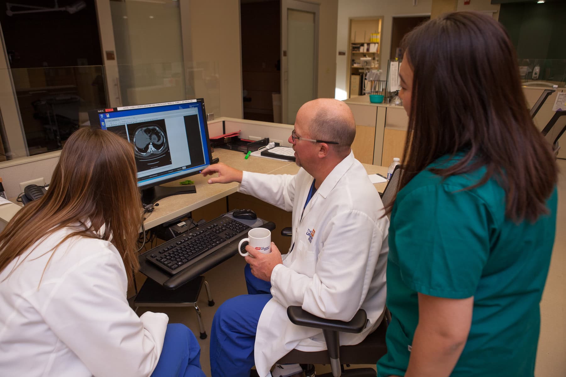 Three medical professionals in the urgency room review a scan on a monitor, with one pointing out details in this hospital office setting.