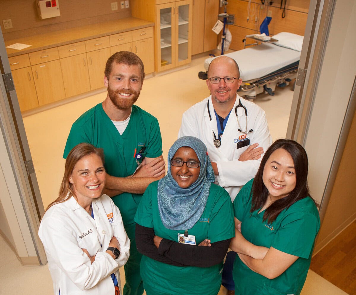 Five healthcare professionals in scrubs and lab coats stand together in the urgency room, smiling at the camera.