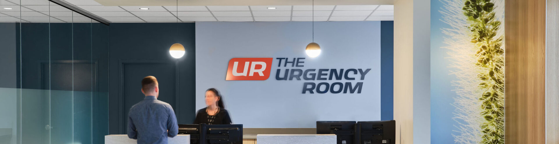 A man stands at The Urgency Room's reception desk, inquiring about an urgent care virtual visit amid modern decor and computers.