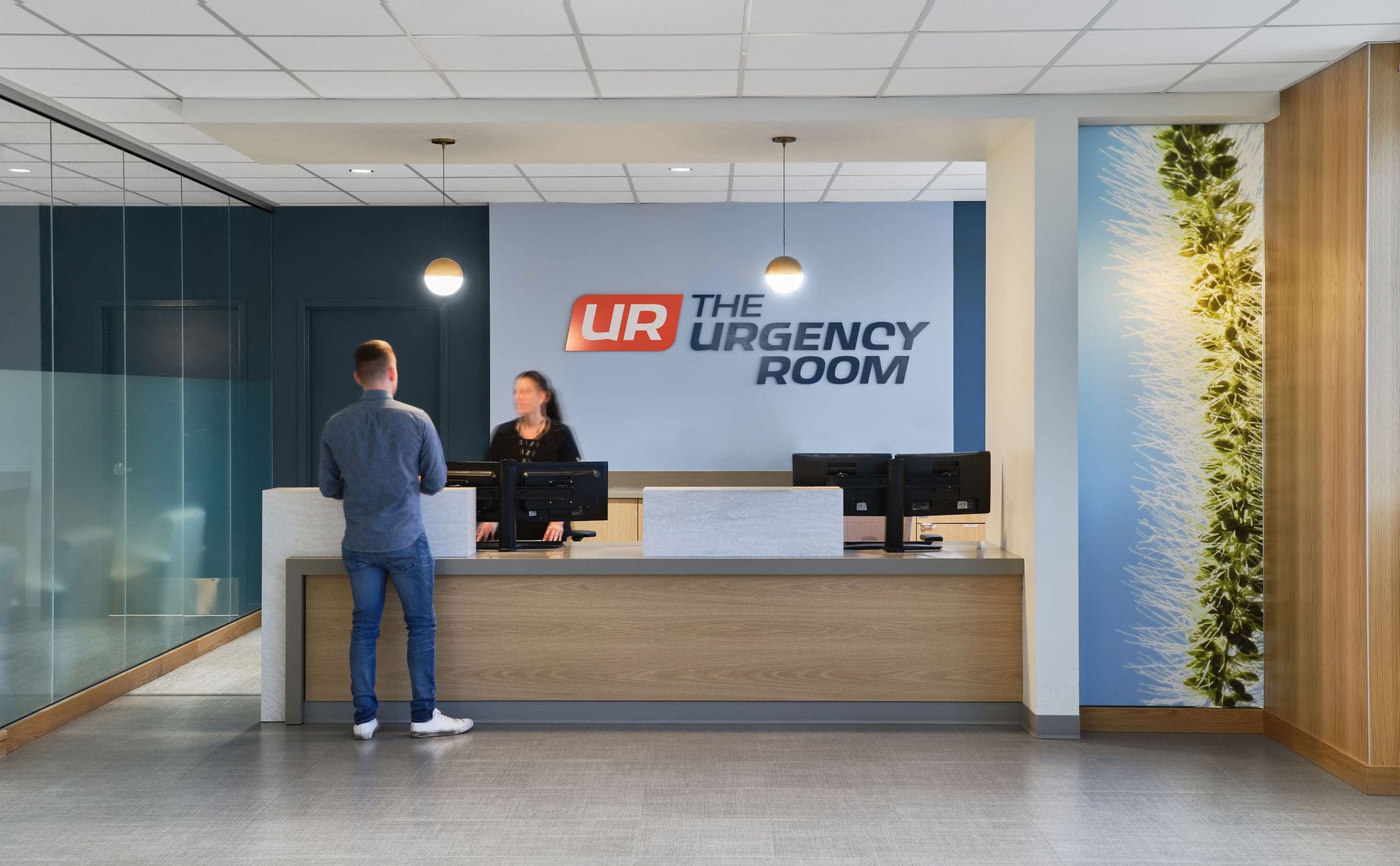 A man stands at The Urgency Room's reception desk, inquiring about an urgent care virtual visit amid modern decor and computers.