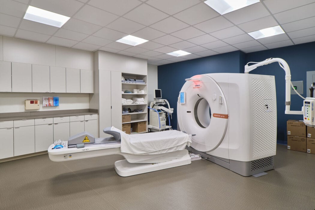 A clean, modern CT scan room at The Urgency Room with a large scanner, exam table, cabinets, and advanced medical equipment.