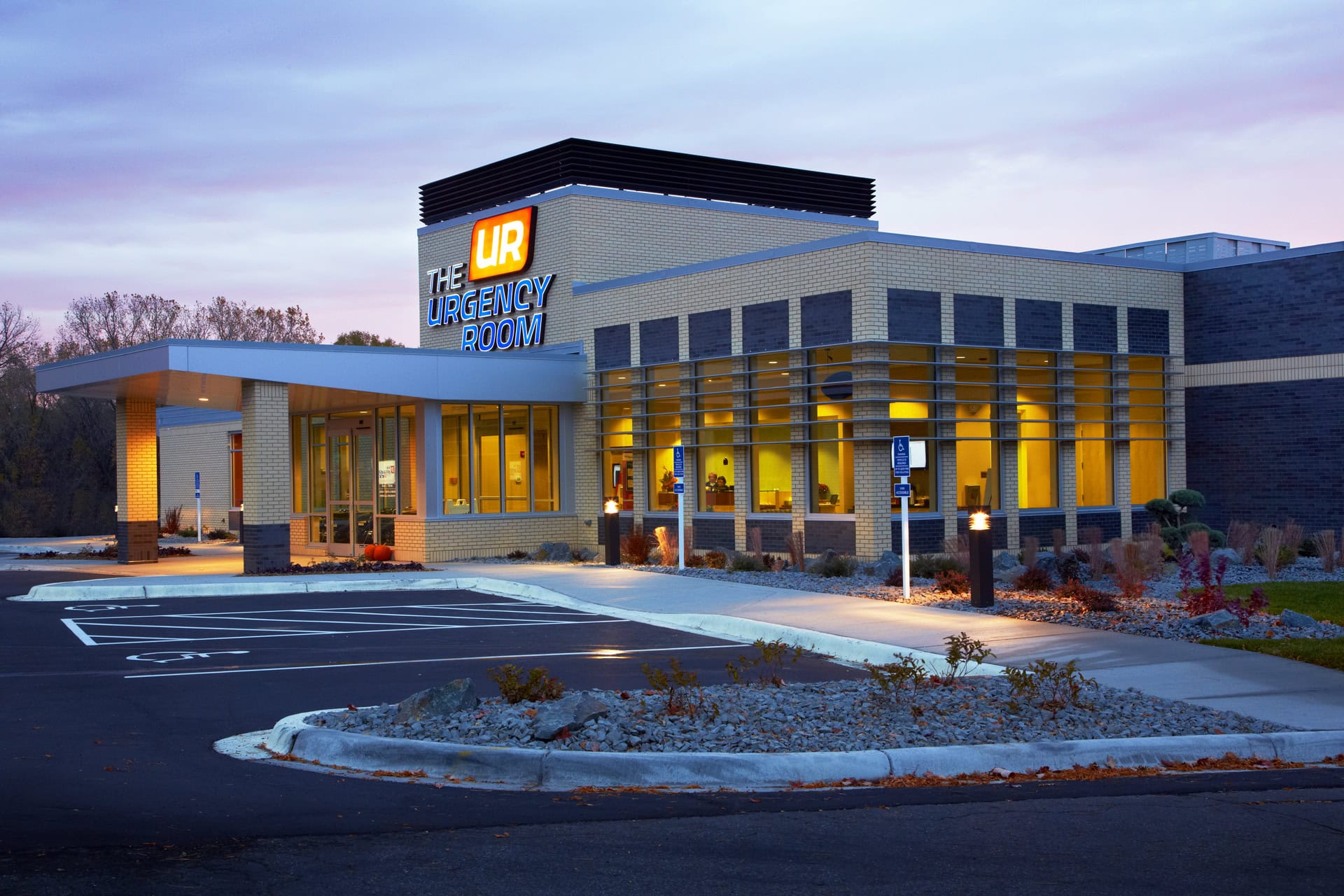 A modern, well-lit building labeled "The Urgency Room" offers online urgent care with large windows and an empty parking lot at dusk.