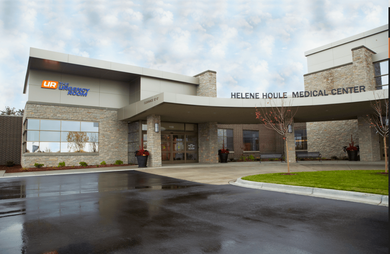 The entrance of Helene Houle Medical Center with “The Urgency Room” sign, glass doors, and wet pavement hints at online urgent care options here.