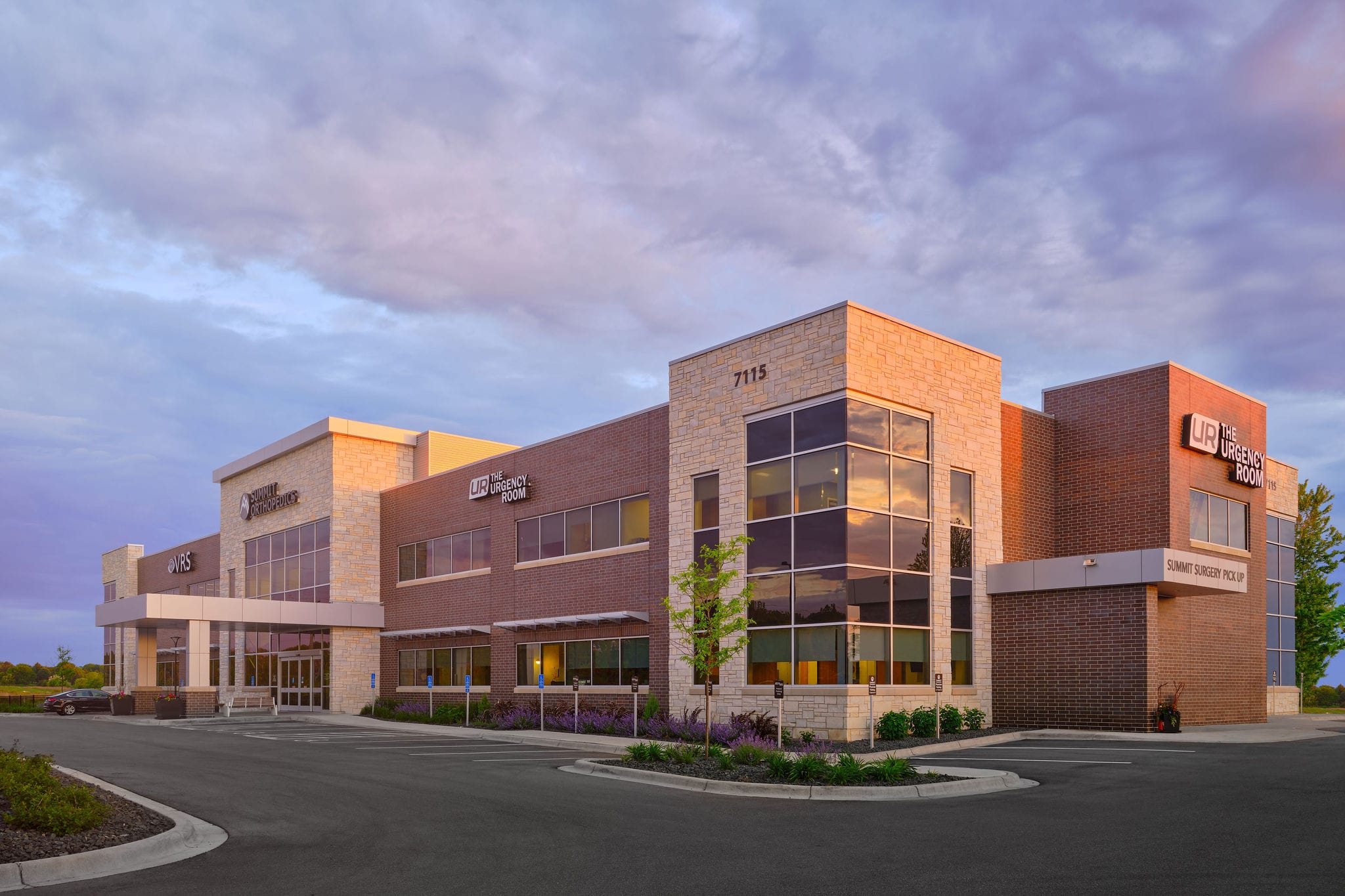 A modern two-story medical office with brick and stone exterior offers online urgent care and ample parking, glowing at sunset.